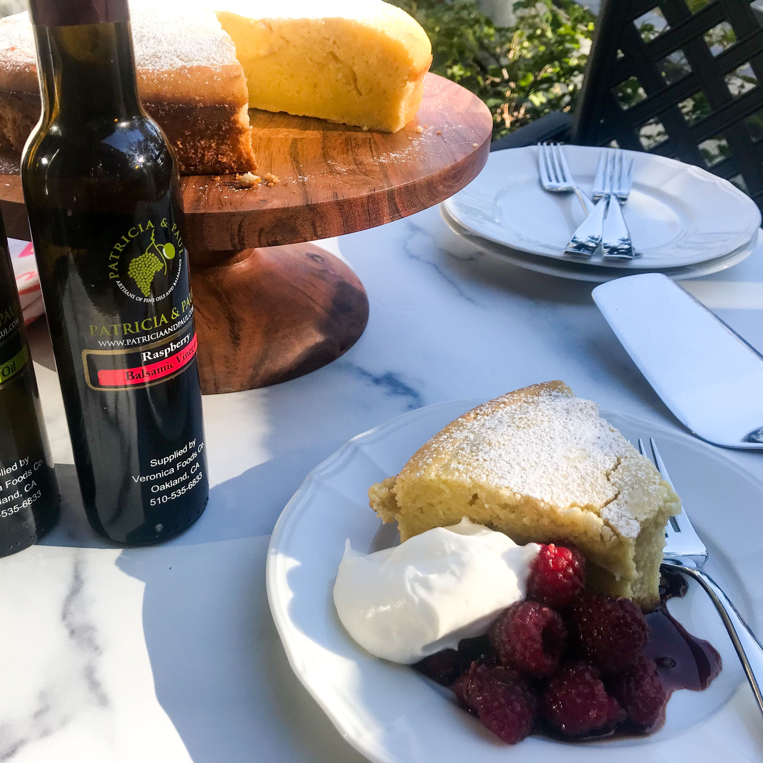 a slice of lemon olive oil cake with balsamic raspberries and whipped cream on a plate with the whole cake and plates and forks on a table.