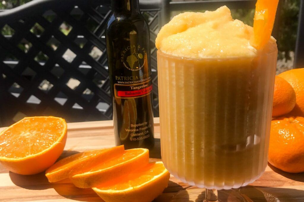 A glass of frozen orange aperol spritz with a bottle of vinegar and fresh oranges