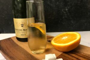 bottle of champagne and a glass with a champagne cocktail on a cutting board with half an orange and sugar cube