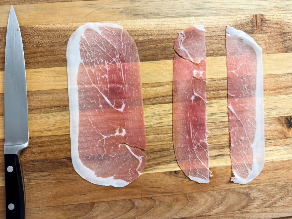 Three slices of ham arranged on a wooden cutting board beside a sharp knife.