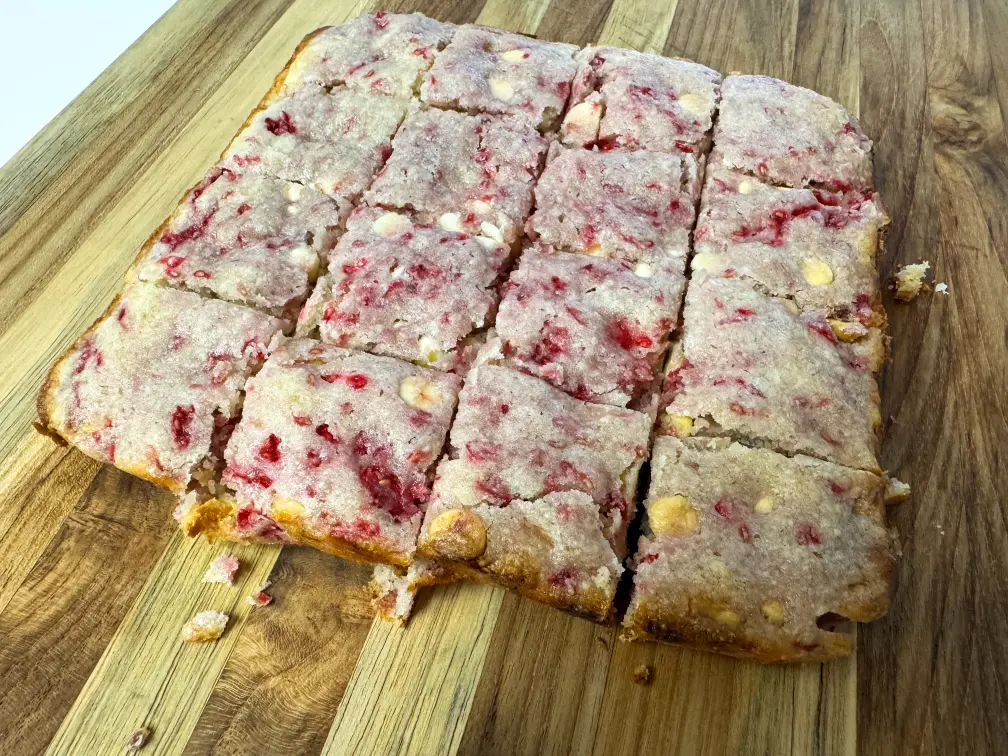 Raspberry white chocolate blondies cut into squares on a wooden board.